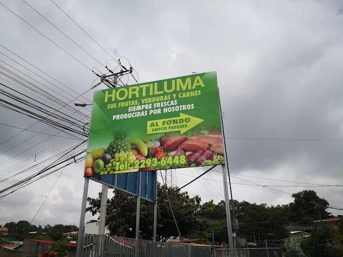 Rótulo de Hortiluma en un poste sobre cables eléctricos: frutas, verduras y carnes, siempre frescas, producidas por nosotros, amplio parqueo al fondo.